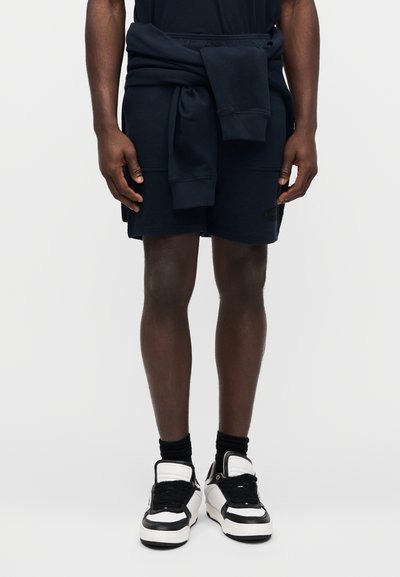 Σκούρα μπλε σορτς με κορδόνι στη μέση, συνδυασμένα με σκουρόχρωμο μπλε φούτερ δεμένο στη μέση. Τα παπούτσια είναι μαύρα και λευκά sneakers με ψηλή κορυφή.