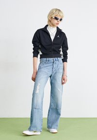 Svart zip-up jacka med vita sidopaneler, slitna ljusblå vida jeans och vita skor. Accessoarer inkluderar solglasögon och smycken.