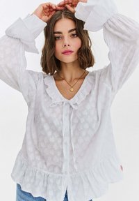 Kobieta o brązowych włosach do ramion ubrana w białą, teksturowaną bluzkę z falbankami i złoty naszyjnik z wisiorkiem, unosi ramiona nad głową.