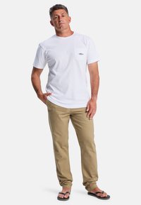 White cotton t-shirt with a chest pocket, paired with beige pants and black flip-flops. Simple design with a relaxed fit.