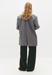 Cappotto grigio in misto lana con una vestibilità rilassata, spalle scese e tasche laterali. Indossato con pantaloni neri a gamba larga, stivaletti texture con tacco basso.