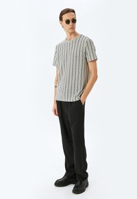 Camiseta de manga corta a rayas en blanco y negro, combinada con pantalones negros y botas negras hasta el tobillo, de pie sobre un fondo sencillo.