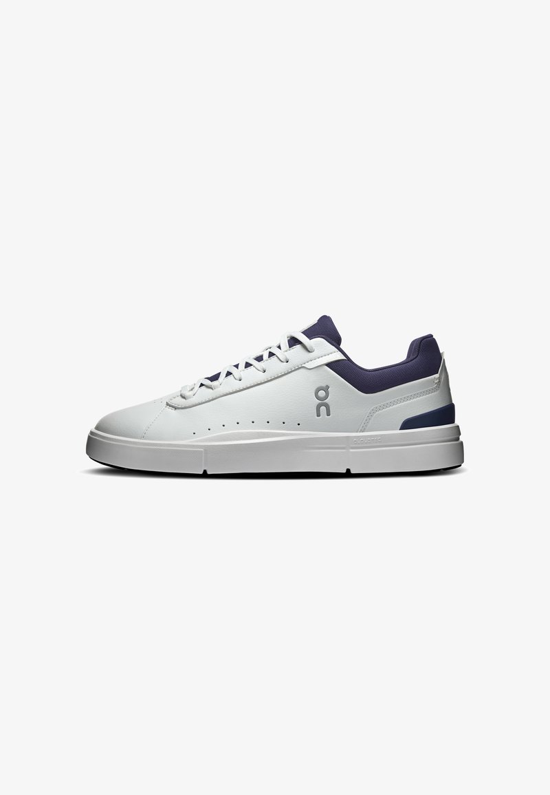 Basket blanche basse avec col et détail du talon bleu marine, fermeture à lacets à l’avant, côté perforé et logo près des lacets sur fond blanc.