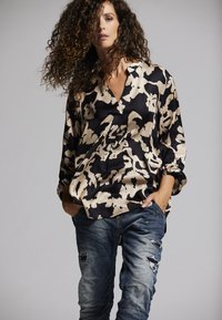 Schwarz-beige gemusterte Bluse mit V-Ausschnitt und langen Ärmeln, kombiniert mit ausgebleichten blauen, zerrissenen Jeans. Lockige Haare prominent zur Schau gestellt.