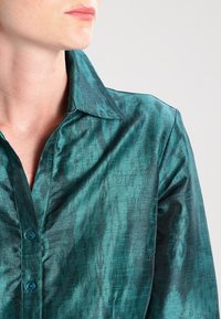 Teal silk shirt featuring a structured collar, front buttons in a contrasting color, and a subtle textured pattern across the fabric.