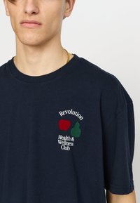 Marineblauw katoenen T-shirt met een ronde hals, korte mouwen en een geborduurd logo met appels, peren en tekst in wit en rood.