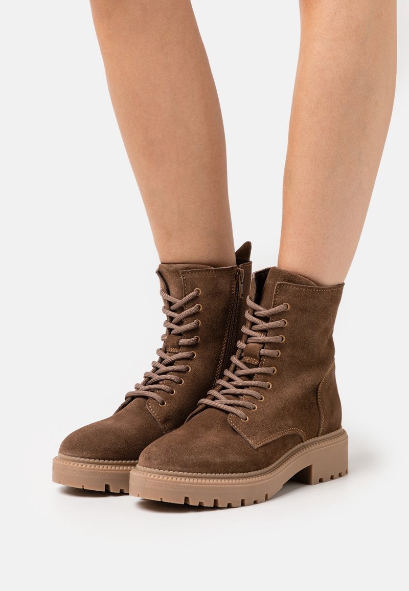 Bottes cheville en daim marron à lacets avec fermeture éclair latérale, portées sur jambes nues, dotées de semelles épaisses en caoutchouc beige avec une adhérence rainurée.