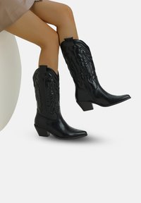 Botas de vaquero negras hechas de cuero suave, con diseños florales en relieve, caña en ángulo y un tacón bajo y cuadrado.