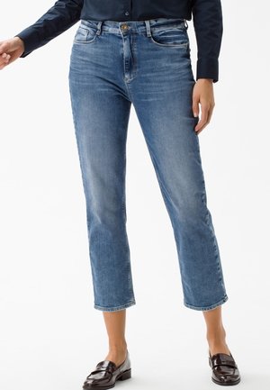 Woman wearing mid-rise, cropped blue jeans and dark loafers, paired with a dark long-sleeve top, standing against a white background.