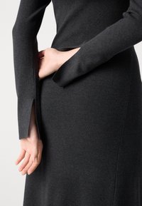 Personne portant une robe en maille gris foncé à manches longues avec des fentes pour les pouces, montrant les mains placées derrière le dos.