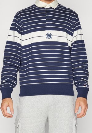 Mężczyzna noszący granatowo-białą pasiastą koszulkę rugby z długim rękawem z logo New York Yankees i białym kołnierzem, w połączeniu z jasnoszarymi spodniami typu cargo.