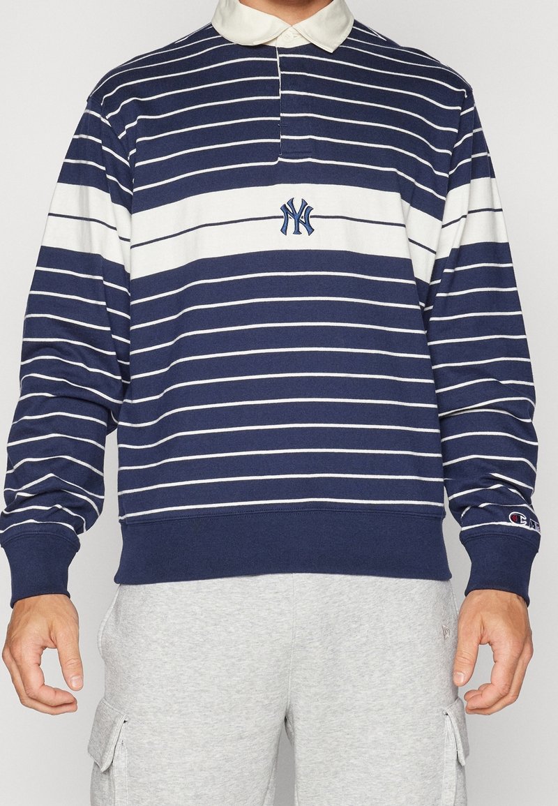Homme portant un maillot de rugby manches longues rayé bleu marine et blanc avec le logo des New York Yankees et un col blanc, associé à un pantalon cargo gris clair.
