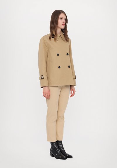 Manteau trench beige à double boutonnage avec des boutons noirs, associé à un pantalon beige ajusté et des bottines noires avec des accents argentés.