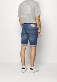 Osoba stojí zády k fotoaparátu, má na sobě šedé tričko a vyhrnuté džíny, doplněné bílými teniskami.