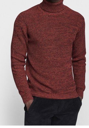 Person trägt einen enganliegenden, weinroten und schwarzen Strickrollkragenpullover und hat die Hände in den Taschen einer dunklen Hose vor einem einfarbigen Hintergrund.