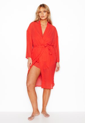 Femme debout pieds nus, portant une robe rouge transparente avec un motif texturé, nouée à la taille, et un short assorti en dessous.