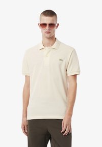 Krämfärgad polotröja med korta ärmar, tvåknapplåsning, broderad grön logotyp på vänster bröst, i kombination med mörka byxor och solglasögon.
