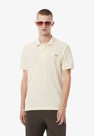 Polo crème à manches courtes, avec patte de boutonnage à deux boutons, logo vert brodé sur la poitrine gauche, associé à un pantalon sombre et des lunettes de soleil.