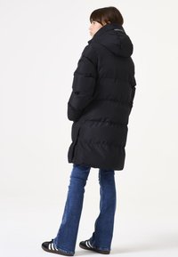 Chaqueta de plumas negra con capucha, con un diseño acolchado y botones de presión en los laterales, combinada con jeans de mezclilla acampanados y zapatillas negras.