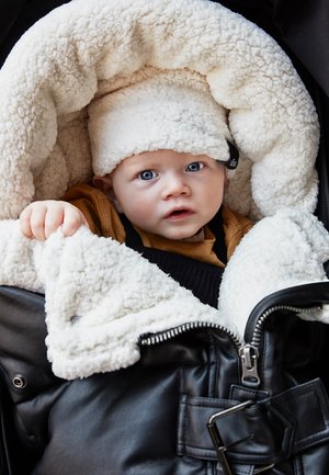 Infant with blue eyes wearing a fluffy white hat and brown outfit, wrapped in a black and white fleece-lined jacket.