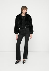 Chaqueta corta de piel sintética negra con un cuello alto, combinada con jeans negros de campana y tacones negros de punta. Textura suave en toda la prenda.