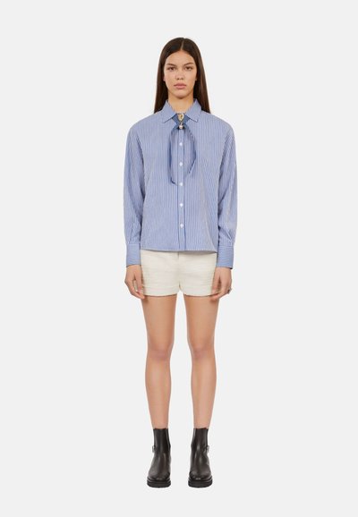 Camicia a righe blu e bianca, con cravatta e maniche lunghe, abbinata a pantaloni corti crema e stivali neri alla caviglia.