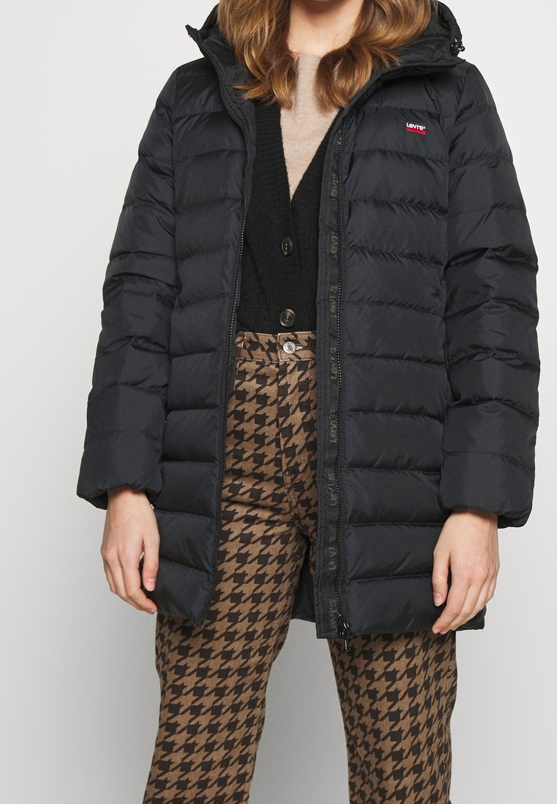 Kobieta ubrana w czarną puchową kurtkę z logo Levi's, czarny kardigan, beżowa bluzka i brązowe spodnie w jodełkę, stojąca na tle jednolitego tła.