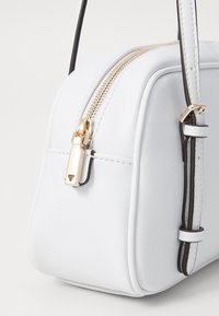 Bolso de cuero sintético blanco con un diseño estructurado, cremallera dorada y delgados acentos en negro. Presenta correas ajustables y una textura suave.
