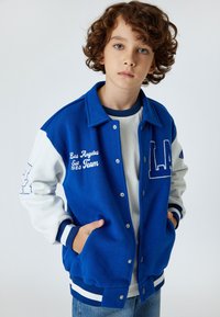 Giacca varsity blu con maniche bianche, con ricamo della scritta "Los Angeles Team" e logo. Polsini e orlo a coste, chiusura con bottoni a pressione.