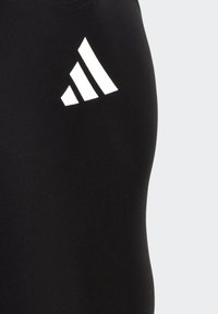 Leggings deportivos negros, fabricados con un material liso y elástico, presentan un logo de tres franjas blancas en la parte superior izquierda.