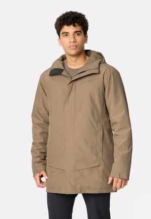 Jonge man met een beige waterdichte jas met capuchon en voorzakken en zwarte broek, staand tegen een effen lichte achtergrond.