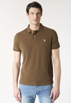 Uomo con polo marrone a maniche corte e jeans azzurri, in piedi davanti a uno sfondo bianco uniforme.