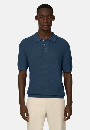 Mężczyzna w niebieskim, krótkim rękawie z dzianiny polo z trzema guzikami i beżowych spodniach, stojący na jasnym, jednolitym tle.