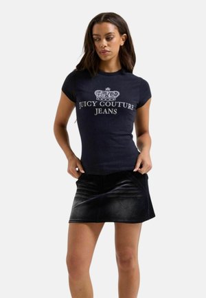 Vrouw draagt een zwart Juicy Couture Jeans T-shirt met een kroongrafiek en een zwarte gestructureerde minirok tegen een effen achtergrond.