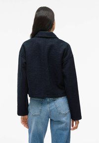 Mörkblå kort jacka i strukturerad tyg, med bred krage och sänkta axlar, tillsammans med ljusblå jeans.