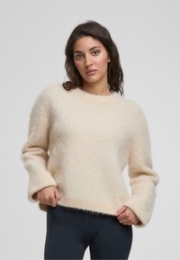 Helles beiges Sweater aus flauschigem Material, mit rundem Ausschnitt, langen Ärmeln und gerippten Bündchen und Saum, sowie einer weichen Textur.