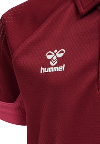 Kastanjebrun sportskjorta med ett texturerat, ventilerande material. Har en vit "hummel"-logotyp och en krage. Kort ärm med accentdetaljer.