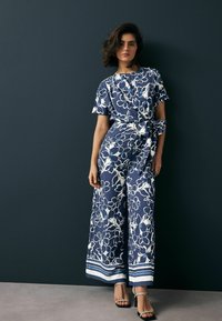 Blommig jumpsuit i marinblått med vita mönster, vida ben, korta ärmar, bälte i midjan och kontrasterande randig fåll. Svarta sandaler.