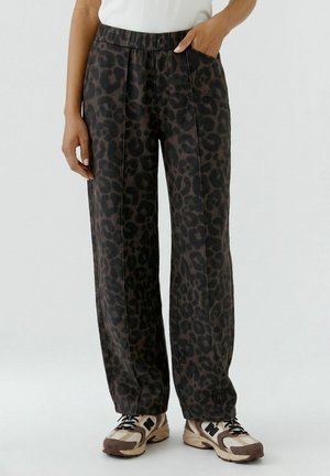Lockere dunkle Hose mit schwarzen Leopardenflecken und sichtbaren vorderen Nähten, getragen mit hellen Sneakers und einem weißen Oberteil.