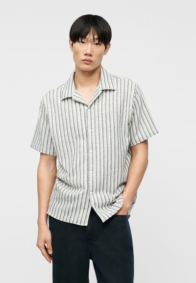 Νέος άνδρας με ίσια μαύρα μαλλιά, φορώντας λευκό πουκάμισο με κοντά μανίκια και κατακόρυφες μαύρες ρίγες και σκούρο παντελόνι, με το χέρι στην τσέπη.