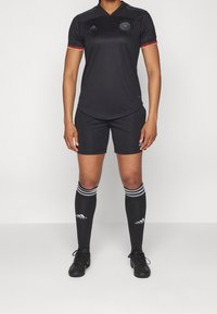 Czarna koszulka piłkarska z krótkim rękawem, logo na piersi, czerwone akcenty, w połączeniu z czarnymi spodenkami, długimi skarpetami do kolan z białymi paskami i korkami.