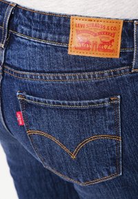 Mörkblå denimjeans med en läderpatch som har röda grafiska tryck, synlig sömnad på bakfickorna och en liten röd logotagg på sidan.