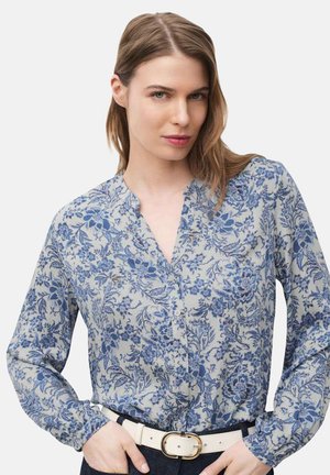 Femme portant un chemisier à manches longues à fleurs bleues et blanches avec une ceinture blanche, debout les mains posées sur les hanches, fond neutre.