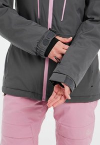 Graue Außensjacke mit rosa Akzenten, strukturiertem Stoff, Klettverschlussmanschetten und tailliertem Design, kombiniert mit hellrosa Hosen mit Cargo-Taschen.