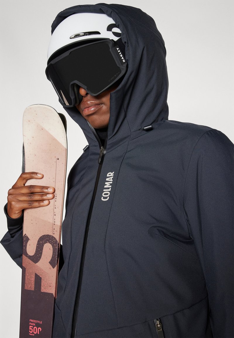 Dunkelblaue Skijacke mit Kapuze und Reißverschlus, die das "Colmar"-Logo trägt. Das Modell hält einen Ski mit einem pinken und schwarzen Design und trägt schwarze Skibrillen.
