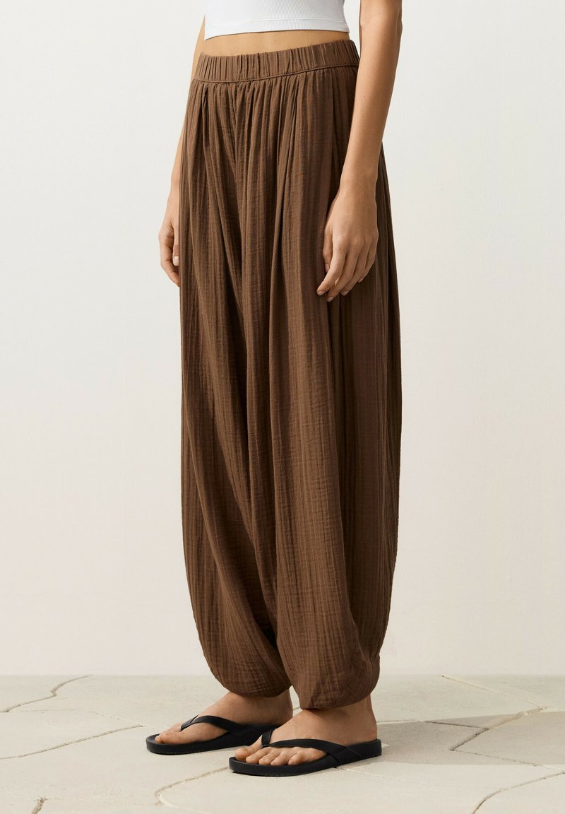 Pantalon ample marron texturé avec taille élastique, assorti à des tongs noires, porté par une personne debout visible de la taille aux pieds.
