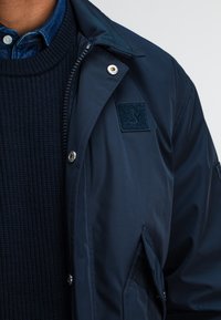 Marineblaue, wasserabweisende Jacke mit glatter Oberfläche, ausgestattet mit einem aufgenähten Logo, einer Reißverschlusstasche und einem geknöpften Kragen über einem strukturierten Pullover.