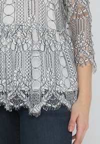 Spetstopp med en genomskinlig vit bas, som har svarta blommönster, volanger vid fållen och texturerade detaljer på ärmarna.