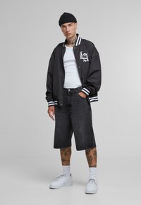 Svart varsity-jacka med vita ränder, vit linne, svarta jeansshorts och vita sneakers. Modellen har tatueringar och en beanie.