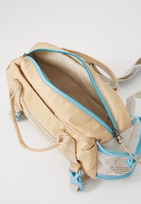 Beige stoffen tas met een lichtblauwe rits, mesh zijkanten en geweven handvatten; heeft een ruime binnenkant voor het dragen van spullen.